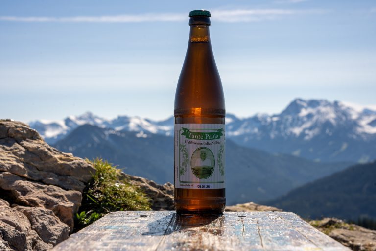 Tante Paula Bierflasche steht auf einer Holzbank auf einem Berggipfel. Im Hintergrund sieht man Bergpanorama.