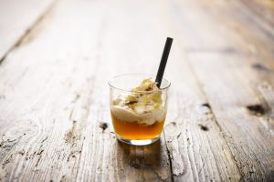 Hausgemachter Bierlikör mit Sahnehaube und kleinem Strohhalm auf einem Holztisch.