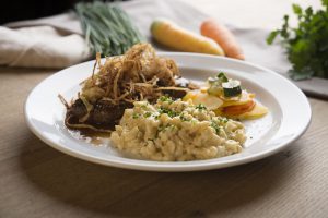 Zwiebelrostbraten mit knusprigen Röstzwiebeln, cremigen Spätzle und buntem Ofengemüse auf einem weißen Teller, angerichtet auf einem Holztisch.