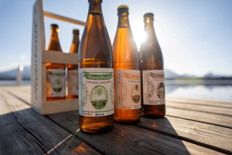 Unsere selbstgebrauten Biere stehen auf einem Steg. Im Hintergrund sieht man den Hopfensee, Berge und Sonne scheint von hinten durch die Bierflaschen.