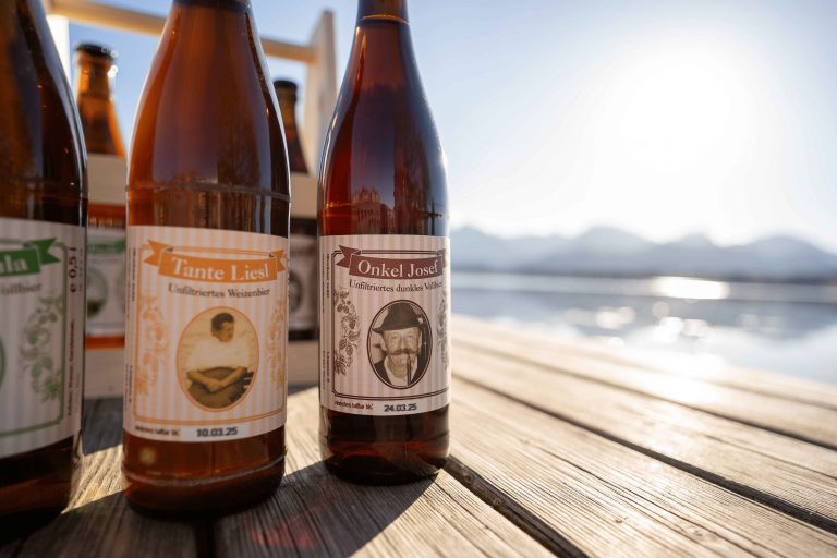 Unsere selbstgebrauten Biere auf einem Holzsteg, im Vordergrund sieht man das Onkel Josef Bier, im Hintergrund sieht man den See und Bergpanorama.
