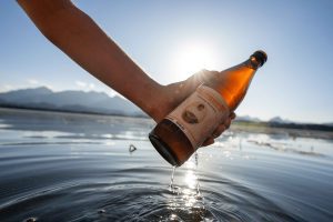 Eine Person hält eine Tante Liesl Bierflasche überhalb des Hopfensees, umgeben von sanften Wellen und Sonnenlicht in Hopfen am See.