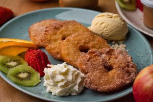 Knusprige Apfelküchle auf einem blauen Teller, mit Vanilleeis, Sahne und frisches Obst.