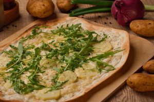 Knuspriger Flammkuchen mit Kartoffelscheiben, Käse und Rucola belegt auf einem Holzbrett serviert.