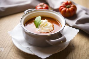 Hausgemachte Tomatensuppe in einer grauen Suppenschale, garniert mit Sahnehäubchen und frischem Basilikum, dekoriert mit frischen Tomaten im Hintergrund.
